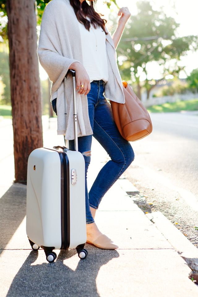 Travel outfit featuring an oatmeal sweater with distressed denim and nude flats, with a white carry-on. As seen on Kendi Everyday