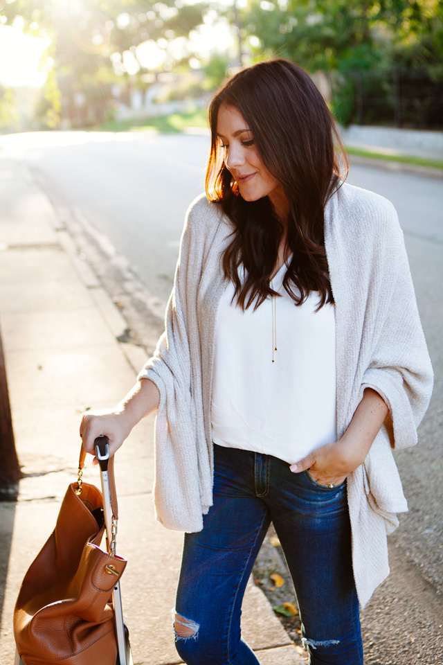 Travel outfit featuring an oatmeal sweater with distressed denim and nude flats, with a white carry-on. As seen on Kendi Everyday