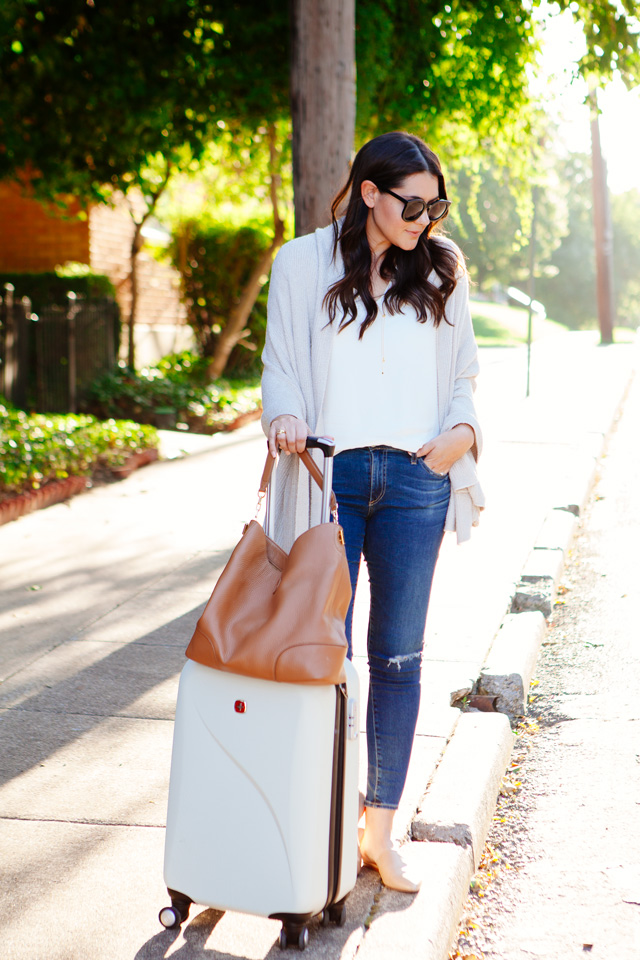 Travel outfit featuring an oatmeal sweater with distressed denim and nude flats, with a white carry-on. As seen on Kendi Everyday