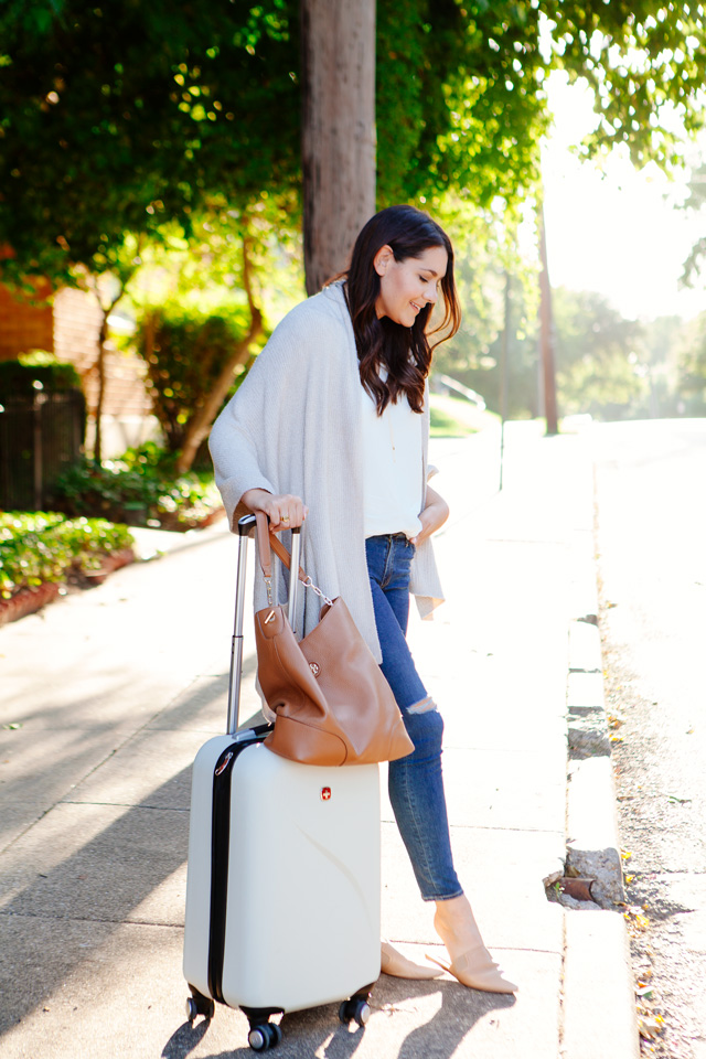 Travel outfit featuring an oatmeal sweater with distressed denim and nude flats, with a white carry-on. As seen on Kendi Everyday